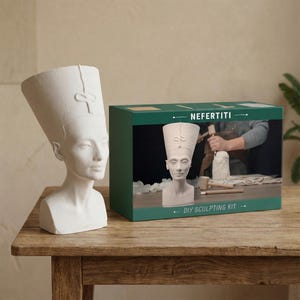 May include: A white bust of Nefertiti stands next to a green box labeled "Nefertiti DIY Sculpting Kit." The bust is on a wooden table. The box shows a person sculpting a bust. The kit includes tools and a bust.