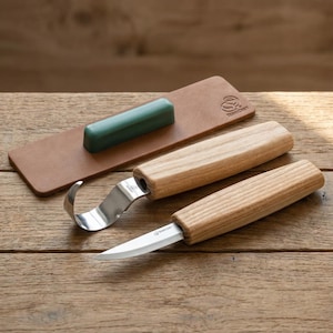 Puede incluir: Un juego de tallado de madera con dos cuchillos, una hoja curva y una hoja recta, y una correa de cuero con un compuesto de afilado verde. Los cuchillos tienen mangos de madera y hojas de acero inoxidable. El juego está sobre una superficie de madera.