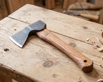 Puede incluir: Un hacha pequeña con mango de madera y cabeza de metal gris. El hacha está sobre una superficie de madera envejecida. El mango tiene un agujero cerca del extremo. Es una herramienta para trabajar la madera.