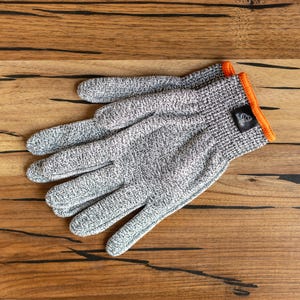 Op de afbeelding: Een paar grijs-witte snijbestendige handschoenen met oranje rand. De handschoenen zijn gemaakt van een textuurrijk materiaal en liggen op een houten oppervlak. Een klein zwart label is zichtbaar op de manchet.