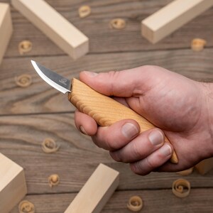May include: A small carving knife with a wooden handle and a curved blade. The knife is held in a hand, with wood shavings and blocks in the background. The blade is silver and the handle is light brown.