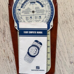 May include: A white flight computer with a blue and white manual. The computer has a circular dial with various scales and markings. The manual has the title "Flight Computer Manual" and the logo "S Sanderson".