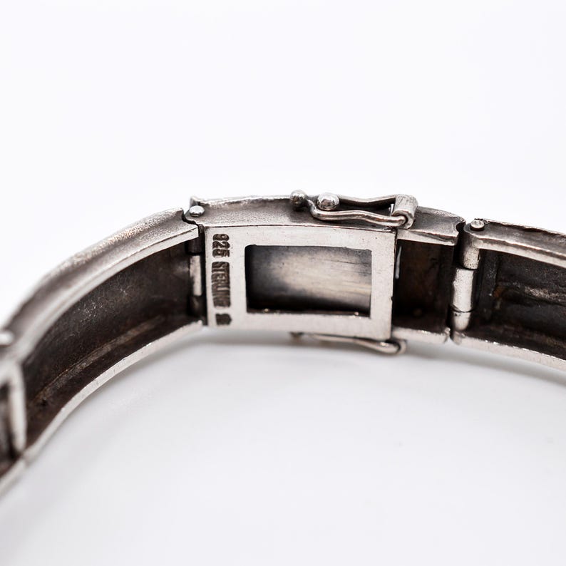 May include: Close-up of a silver bracelet clasp. The bracelet is made of rectangular links and features a secure clasp mechanism. The metal is stamped with "925 STERLING". The bracelet is on a white background.