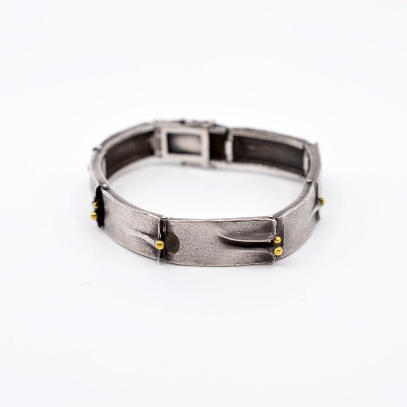 May include: A silver-toned bracelet with a textured surface and a rectangular clasp. The bracelet features gold-colored accents and a unique design. The bracelet is shown on a white background.