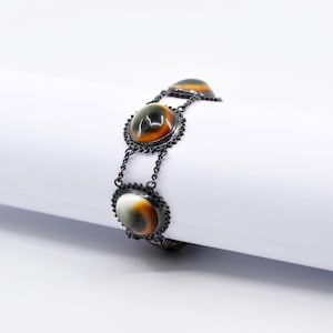 May include: A silver bracelet with three oval-shaped stones set in a black metal frame. The stones are a mix of brown, white, and black.