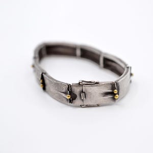 May include: A silver-toned bracelet with a rectangular design. The bracelet has a clasp closure and is accented with small gold-colored beads. The bracelet is shown on a white background.