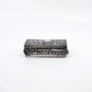 K&ouml;nnte beinhalten: Eine rechteckige Silberdose mit einem verzierten Blumenmuster. Die Dose hat einen Klappdeckel und ist mit filigranen Details verziert.