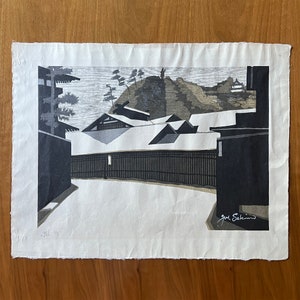 May include: A black and white woodblock print of a Japanese village scene. The print depicts a group of buildings with snow-covered roofs and a hill in the background. The artist's signature, "Seiho", is visible in the lower right corner.
