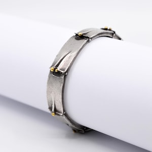 May include: A silver-toned bracelet with a textured, hammered appearance. The bracelet features rectangular links with small gold-colored accents. The design is modern and minimalist, suitable for everyday wear.