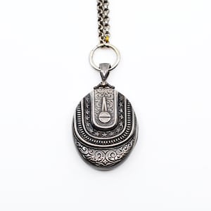 May include: A silver locket necklace with an oval-shaped pendant featuring intricate engraved floral designs. The locket has a hinged closure and a detailed design around the edge.