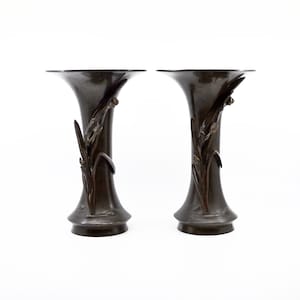 May include: Two bronze-colored vases with a flared top and a detailed floral design. The vases have a slender, elongated shape and are placed side by side against a white background. The floral design appears to be a stylized depiction of reeds or grasses.
