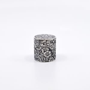May include: A silver cylindrical box with a floral design. The box is embossed with a repeating pattern of flowers and leaves.