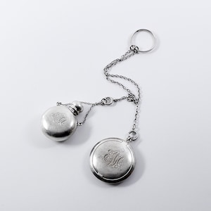 May include: A silver-toned antique accessory featuring a small flask and a round compact, both with engraved initials. The flask has a small screw-on cap. The compact and flask are connected by a delicate chain, with a ring at the top.