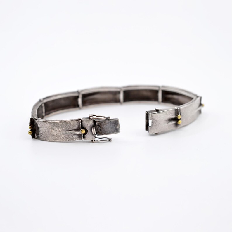 May include: A silver-toned bracelet with a textured finish and an open clasp. The bracelet is composed of rectangular links and features small gold-colored accents. The clasp is open, displaying the inner workings of the jewelry.