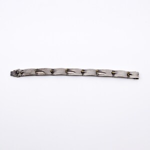May include: A silver-toned bracelet with a series of rectangular links. Each link has a textured surface and is connected by small gold-colored accents. The bracelet is laid flat against a white background.