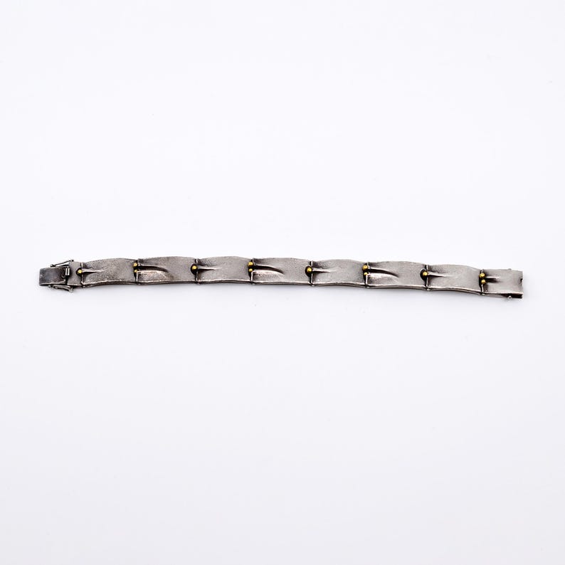 May include: A silver-toned bracelet with a series of rectangular links. Each link has a textured surface and is connected by small gold-colored accents. The bracelet is laid flat against a white background.