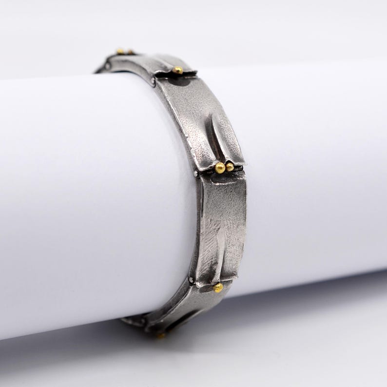 May include: A silver-toned bracelet with a textured surface and gold-colored accents. The bracelet is composed of rectangular links, each featuring a unique design. Small, round gold beads are placed at the joints, adding a touch of contrast to the piece.