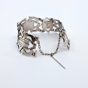 May include: Silver bracelet with an intricate design and a chain closure. The bracelet is marked "Tacco 925".