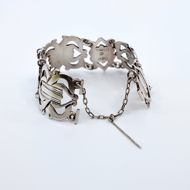 May include: Silver bracelet with an intricate design and a chain closure. The bracelet is marked "Tacco 925".