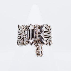 May include: A silver bracelet with an intricate design and a chain detail. The bracelet is open and has a clasp.