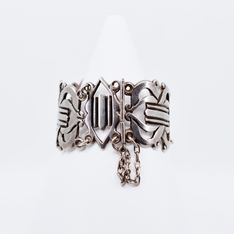 May include: A silver bracelet with an intricate design and a chain detail. The bracelet is open and has a clasp.