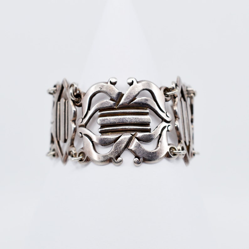 May include: A silver bracelet with an intricate design of interlocking loops and squares. The bracelet is made of a single piece of metal and has a polished finish.