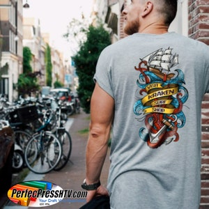 May include: Gray t-shirt with a colorful graphic on the back featuring a ship, anchor, and tentacles. The design includes the text "VISIT KRAKEN SURF SHOP" on a yellow banner. The shirt is being worn by a person walking down a street.