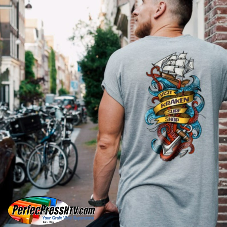 May include: Gray t-shirt with a colorful graphic on the back featuring a ship, anchor, and tentacles. The design includes the text "VISIT KRAKEN SURF SHOP" on a yellow banner. The shirt is being worn by a person walking down a street.
