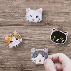 May include: Four embroidered iron-on patches featuring different cat faces. The patches are white with black outlines and feature different coloured eyes and whiskers. The cats are all smiling.
