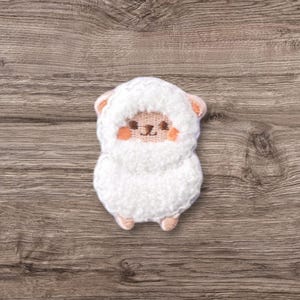 以下が含まれることがあります： かわいい刺繍の顔をした、ふわふわの白い羊のぬいぐるみ。羊はピンクの鼻と頬、黒い目があります。