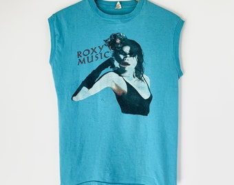 May include: A light blue sleeveless t-shirt with a graphic print of a woman with a flower in her hair. The text "ROXY MUSIC" is printed in red on the shirt.
