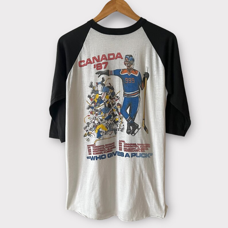 K&ouml;nnte beinhalten: Ein wei&szlig;es und schwarzes Baseball-T-Shirt mit einer Cartoon-Illustration eines Hockeyspielers und dem Text "Canada '87" und "Who Gives A Puck".