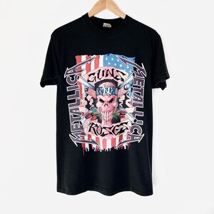 Pode incluir: Camiseta preta com uma estampa gráfica de uma caveira com armas cruzadas e rosas. A caveira está usando uma bandana com o texto "N.N.C." nela. O gráfico é cercado pelo texto "Metallica" e "Guns N' Roses".