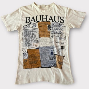 May include: A white t-shirt featuring a collage of printed text and images. The text includes the band name "BAHAUS" and lyrics from their songs. The images include a drawing of a bird and a person's face.
