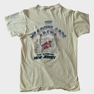 May include: Cream-colored t-shirt with text and graphics. The back of the shirt features the words "The Meadowlands" above a list of dates in July, with "SOLD OUT" in red. The location "East Rutherford, New Jersey" is also printed.