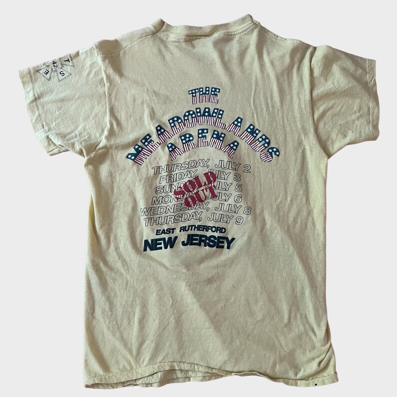 May include: Cream-colored t-shirt with text and graphics. The back of the shirt features the words "The Meadowlands" above a list of dates in July, with "SOLD OUT" in red. The location "East Rutherford, New Jersey" is also printed.