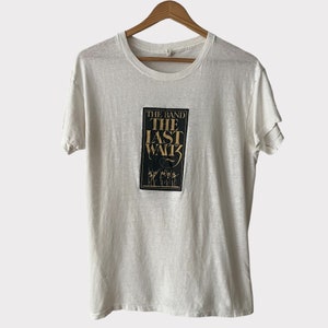 May include: White t-shirt with a graphic from "The Band: The Last Waltz." The graphic is a rectangular design with the text "THE BAND" above "THE LAST WALTZ" in gold lettering. Below the text is a silhouette of musicians. The shirt is hanging on a wooden hanger.