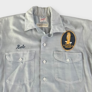 Puede incluir: Camisa de trabajo azul claro con dos bolsillos en el pecho y botones. Presenta el nombre bordado "Bob" y un parche "Richfield" con un diseño de águila. La camisa tiene cuello y una etiqueta pequeña.