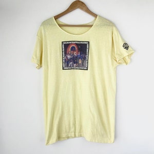 May include: A yellow t-shirt with a graphic of the album cover for "Graham Central Station" by Graham Central Station. The album cover features a group of people dancing in a club with a rainbow above them. The text on the album cover reads "Now Do U Wanna Dance" and "How Do U Wanna Dance".
