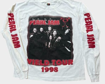 May include: A white long-sleeve t-shirt featuring a black and white photograph of the band Pearl Jam on the front. The text "PEARL JAM", "YIELD TOUR", and "1998" are printed in red. The words "PEARL JAM" are also printed in red down both sleeves.