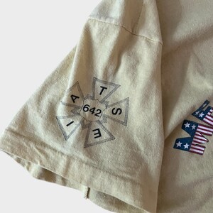 May include: A light yellow t-shirt with a graphic on the sleeve. The graphic is a black and white cross-shaped design with the numbers "642" in the center. The letters "A, T, S, E, I" are around the numbers. The shirt also has a partial American flag design.