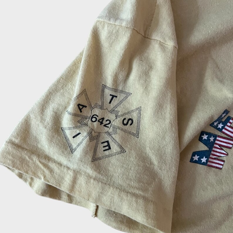 May include: A light yellow t-shirt with a graphic on the sleeve. The graphic is a black and white cross-shaped design with the numbers "642" in the center. The letters "A, T, S, E, I" are around the numbers. The shirt also has a partial American flag design.