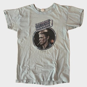 May include: A light blue t-shirt with a vintage David Bowie "On Stage" graphic featuring a black and white image of Bowie with a cigarette in his mouth. The graphic is in a circle with a silver border.