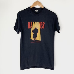 May include: Black t-shirt with a red and yellow graphic of the band Ramones. The graphic features a silhouette of a person in front of a yellow rectangle with the band's name above it and the words "Pleasant Dreams" below it.