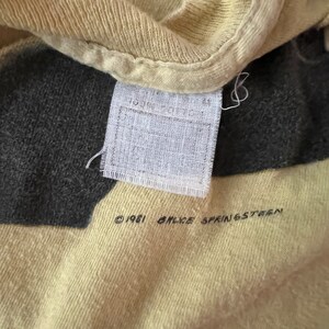 May include: Close-up of a vintage t-shirt with a tag. The shirt is a light yellow color with a dark gray design. The tag reads "100% cotton" and "&copy; 1981 BRUCE SPRINGSTEEN".