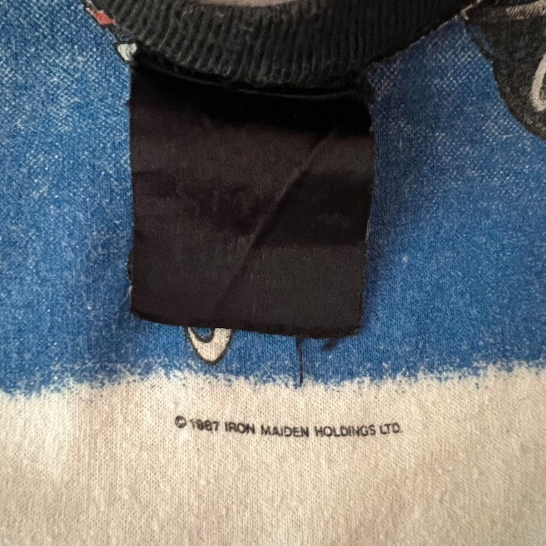 K&ouml;nnte beinhalten: Ein blau-wei&szlig; gestreiftes T-Shirt mit einem schwarzen Band um den Hals. Das Shirt hat einen schwarzen Patch mit dem Wort "Iron Maiden" darauf. Das Shirt hat auch den Text "&copy; 1987 Iron Maiden Holdings Ltd."