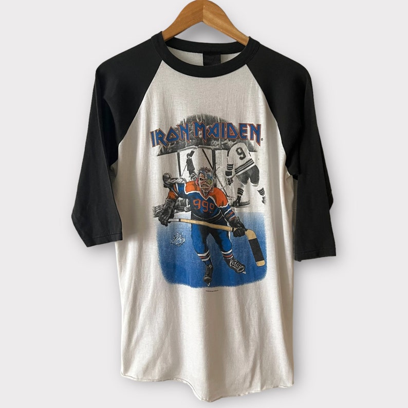 K&ouml;nnte beinhalten: Ein wei&szlig;es und schwarzes Baseball-T-Shirt mit einem Aufdruck eines Hockeyspielers, der ein blaues und oranges Trikot mit der Nummer 999 tr&auml;gt. Der Aufdruck enth&auml;lt auch den Text "IRON MAIDEN" und das Jahr "1986".