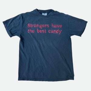 May include: Dark blue t-shirt with the text "Strangers have the best candy" in red. The shirt has short sleeves and a crew neck. The shirt is made of a soft, worn material.