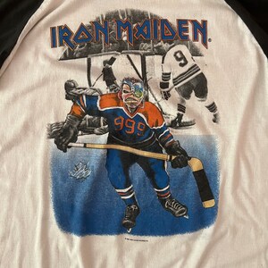 K&ouml;nnte beinhalten: Ein wei&szlig;es T-Shirt mit einem Schwarzwei&szlig;bild eines Hockeyspielers, der ein blaues und orangefarbenes Trikot mit der Nummer 999 auf der Vorderseite tr&auml;gt. Der Text "IRON MAIDEN" ist in Schwarz oben auf dem Shirt gedruckt.