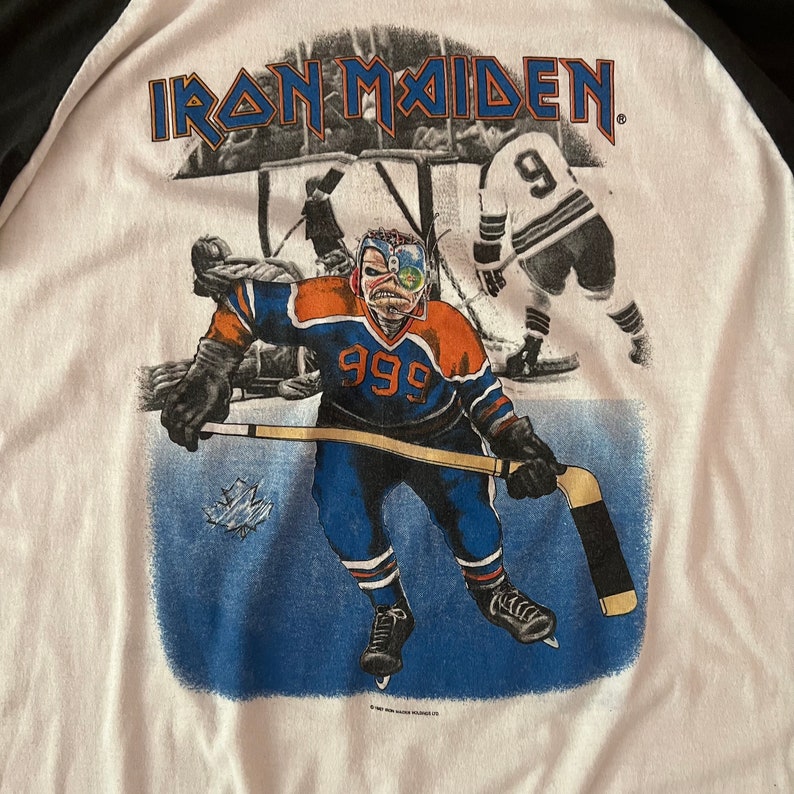 K&ouml;nnte beinhalten: Ein wei&szlig;es T-Shirt mit einem Schwarzwei&szlig;bild eines Hockeyspielers, der ein blaues und orangefarbenes Trikot mit der Nummer 999 auf der Vorderseite tr&auml;gt. Der Text "IRON MAIDEN" ist in Schwarz oben auf dem Shirt gedruckt.